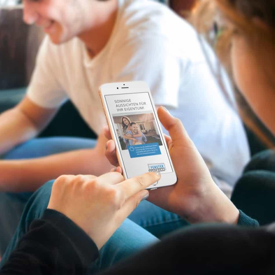 Person hält Smartphone mit digitalem Förderflyer von Fensterbau Hartmann, zeigt Slogan und Bild eines Paares.