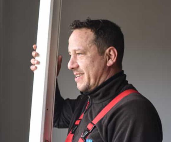 Fenstermonteur von Fensterbau Hartmann inspiziert einen Fensterrahmen, trägt schwarze Arbeitsjacke mit Firmenlogo und rote Sicherheitsträger.