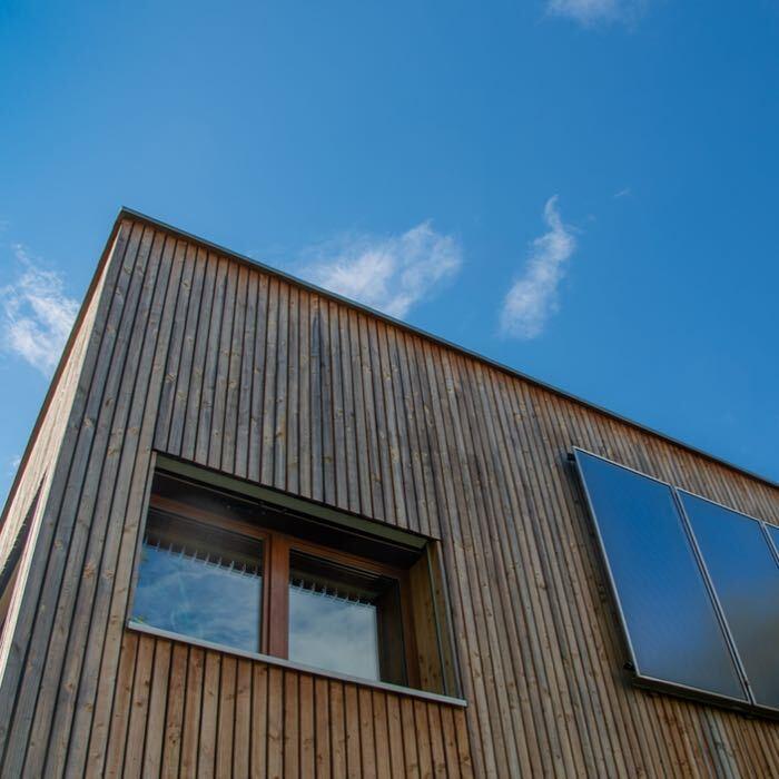Blick Richtung Himmel auf ein Holzhaus mit Holzfenstern und Solar-Panelen.