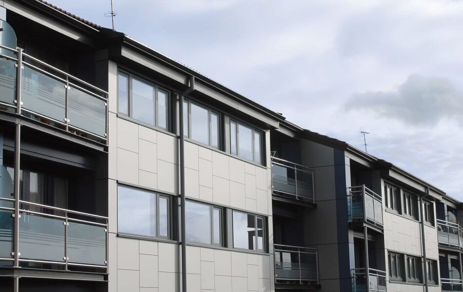 Modernes Mehrfamilienhaus mit grauer Fassade, großen Fenstern und Balkonen mit Glasgeländern. Das Gebäude hat eine zeitgemäße Architektur und klare Linien.