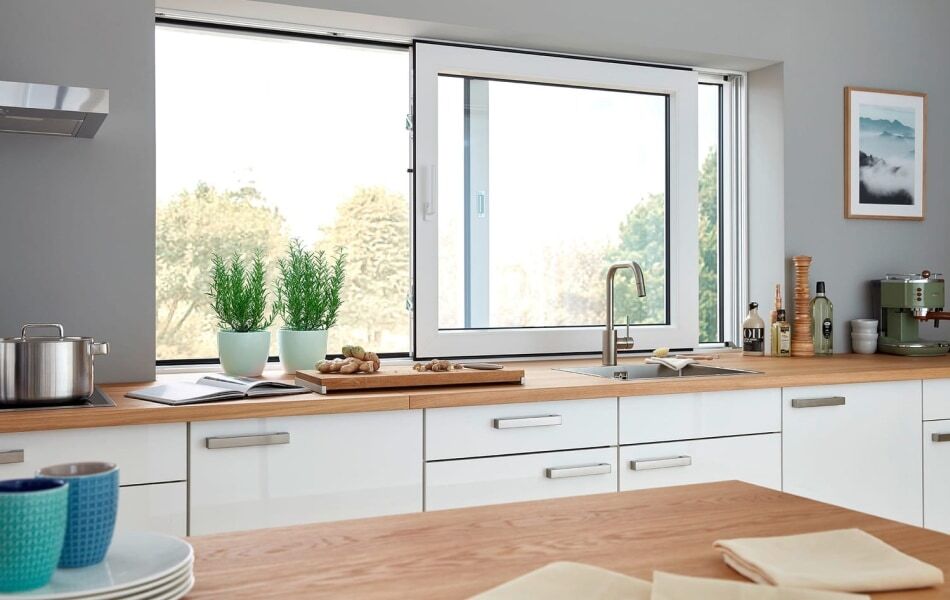Moderne Küche mit weißer Einbauküche, Holzarbeitsplatte und großem Dreh-Kipp-Fenster über der Spüle. Durch das Fenster fällt Tageslicht und bietet Blick ins Grüne.