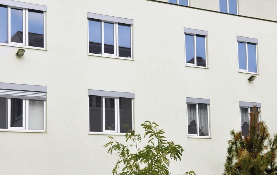 Mehrfamilienhaus mit heller Fassade und modernen Kunststofffenstern in Weiß, teils geöffnet, teils geschlossen. Im Vordergrund sind Bäume zu sehen.